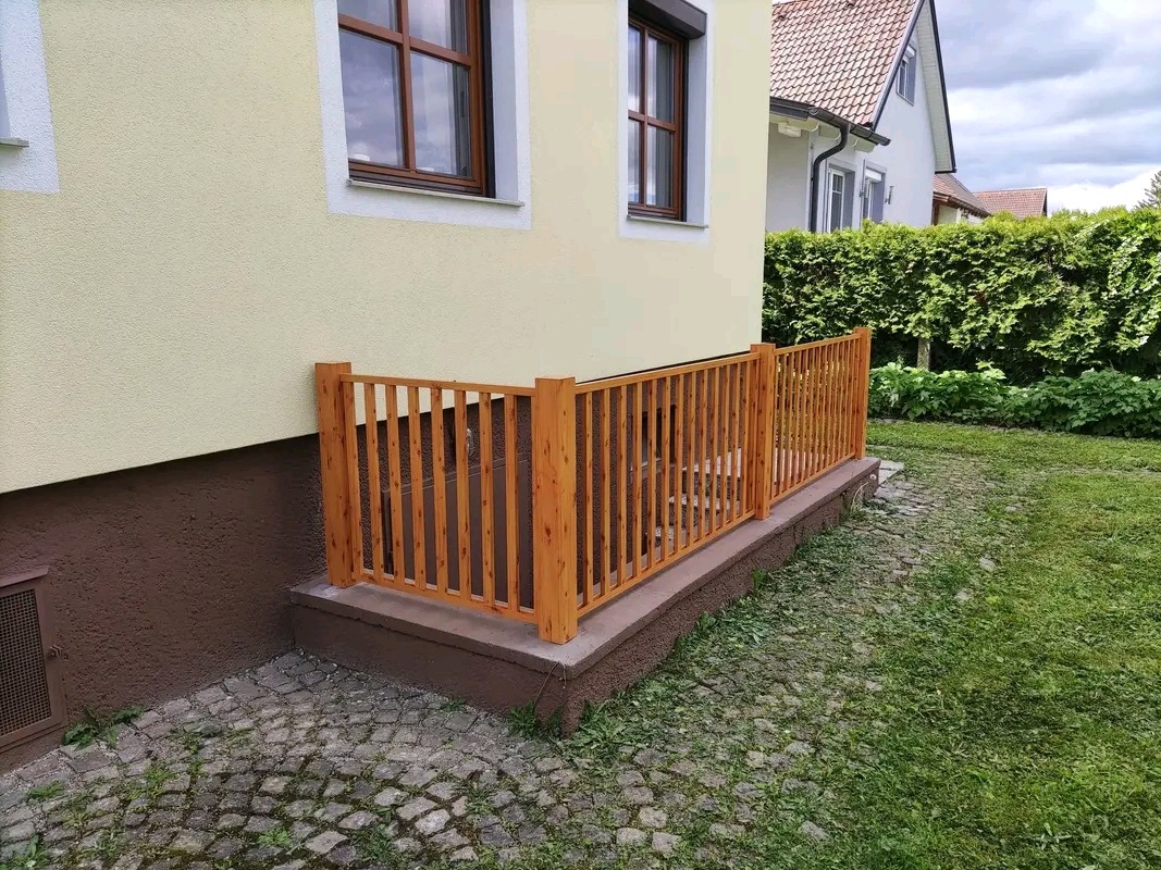 Drewniane ogrodzenie zabezpieczające zejście do piwnicy w domu jednorodzinnym, widoczne z zewnątrz, z trawnikiem i kostką brukową wokół.