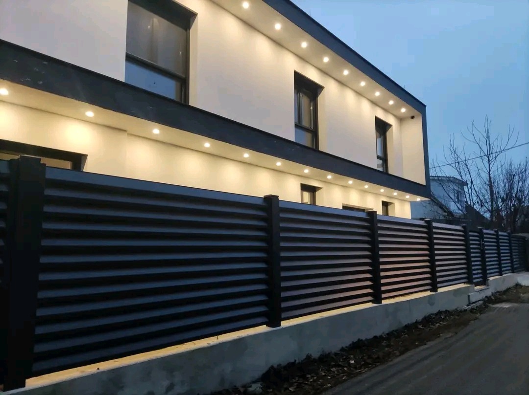 Nowoczesne, poziome ogrodzenie z ciemnych paneli, oświetlony budynek w tle, widok z poziomu ulicy.