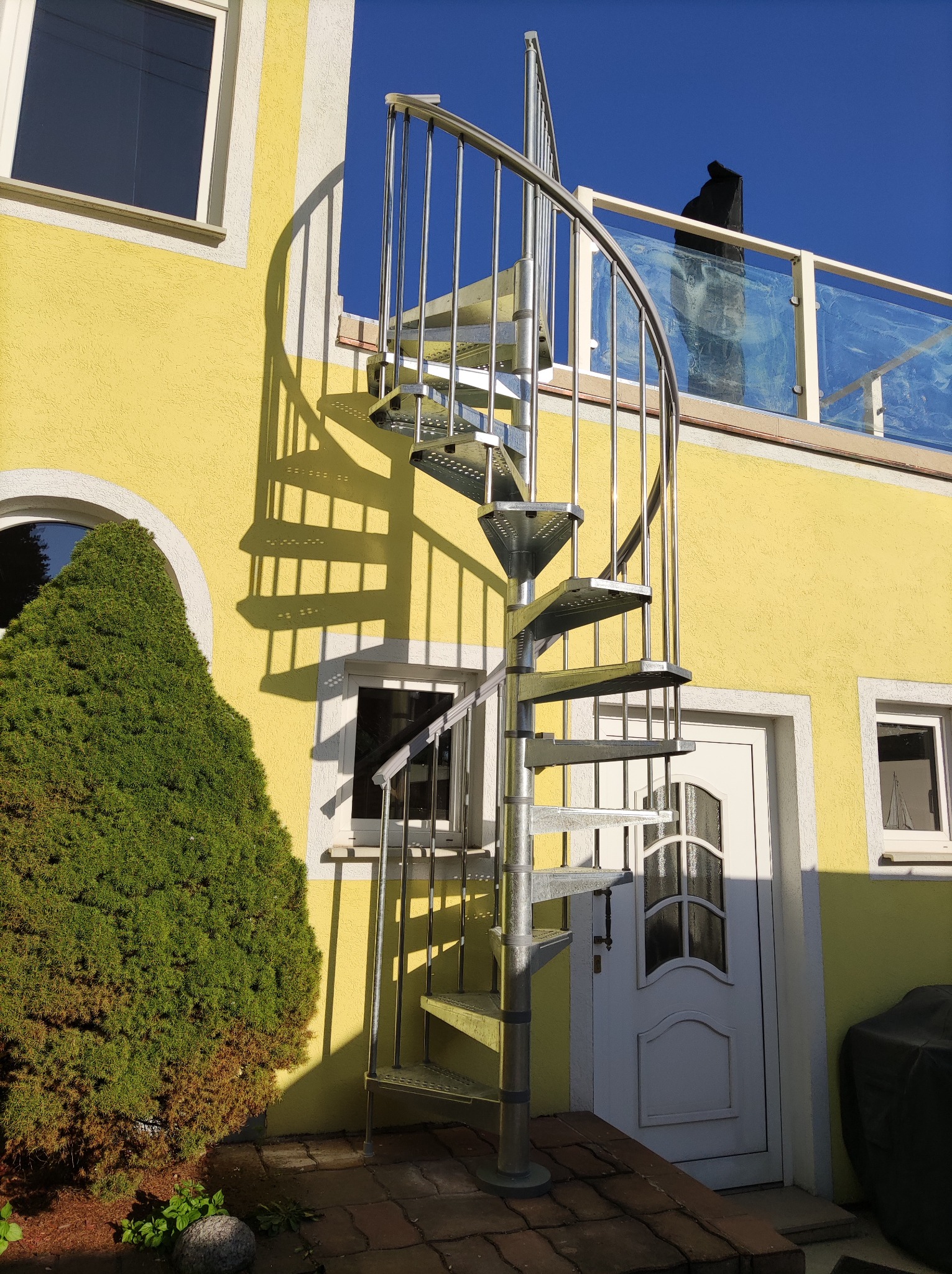 Zewnętrzne schody spiralne z metalu prowadzące na balkon z przeszkloną balustradą, na tle żółtej ściany domu z oknami i białymi obramowaniami.