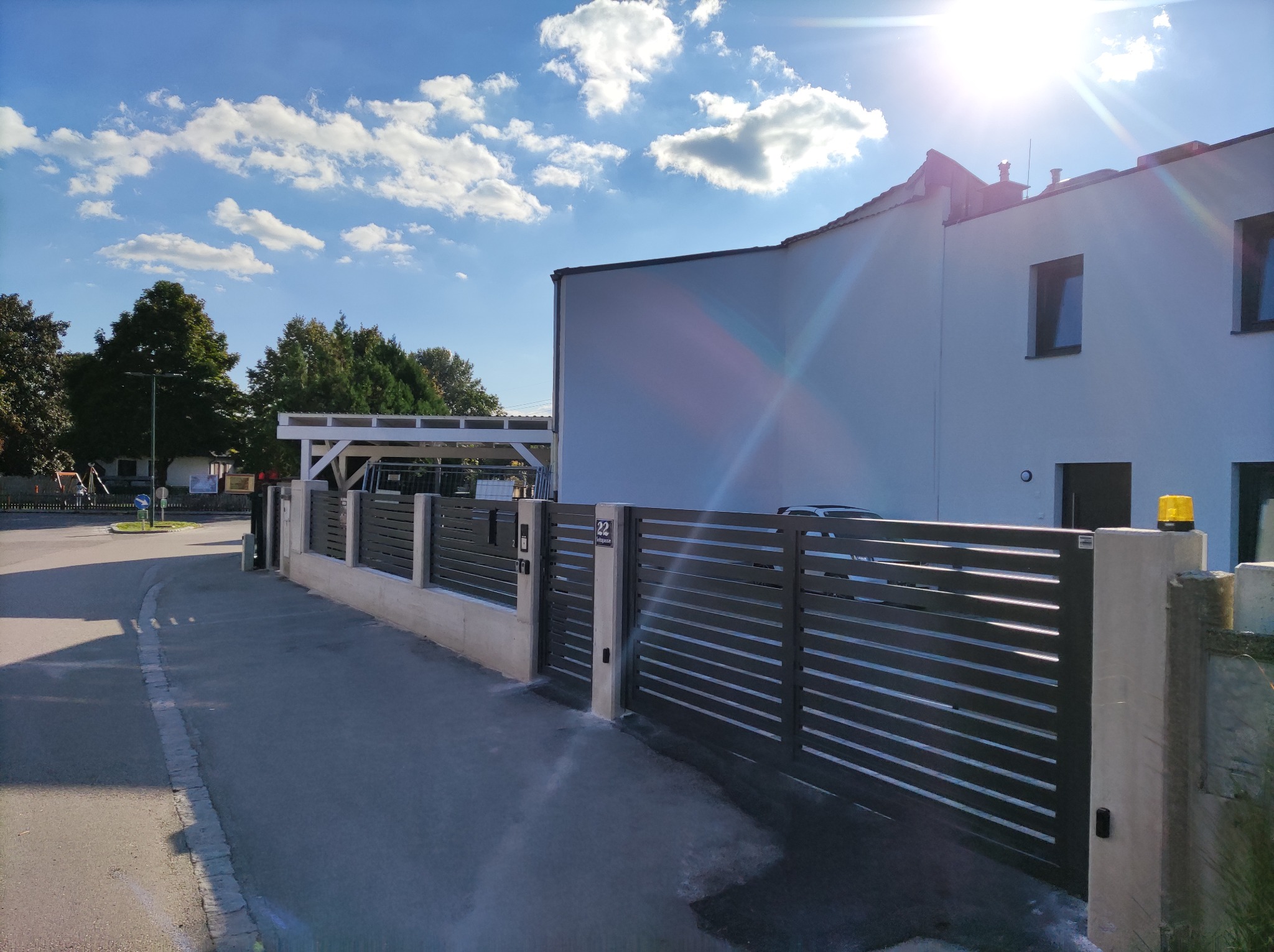 Nowoczesne, poziome przęsła ogrodzenia z metalowymi lamelami i betonowymi słupkami, przed domem z białymi ścianami i niebieskim niebem z chmurami w tle.