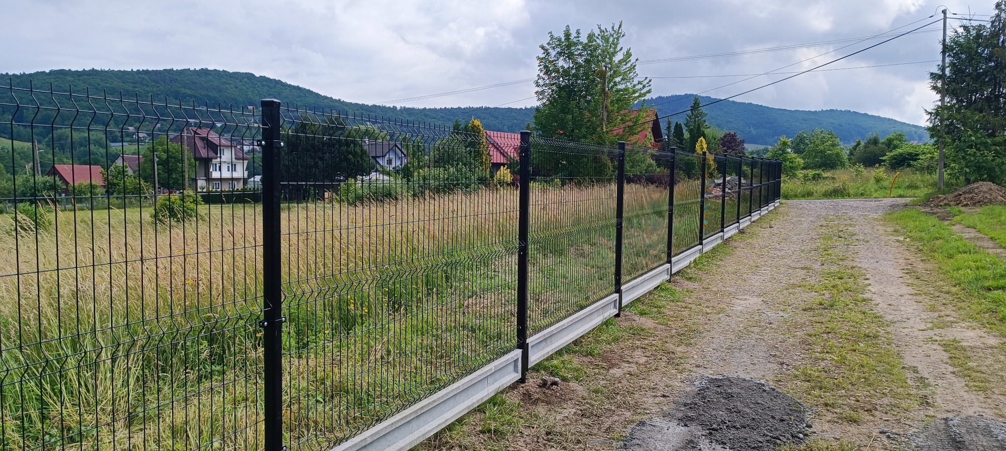 Nowoczesne ogrodzenie panelowe z podmurówką betonową na nierównym terenie, widok na pagórkowaty krajobraz.