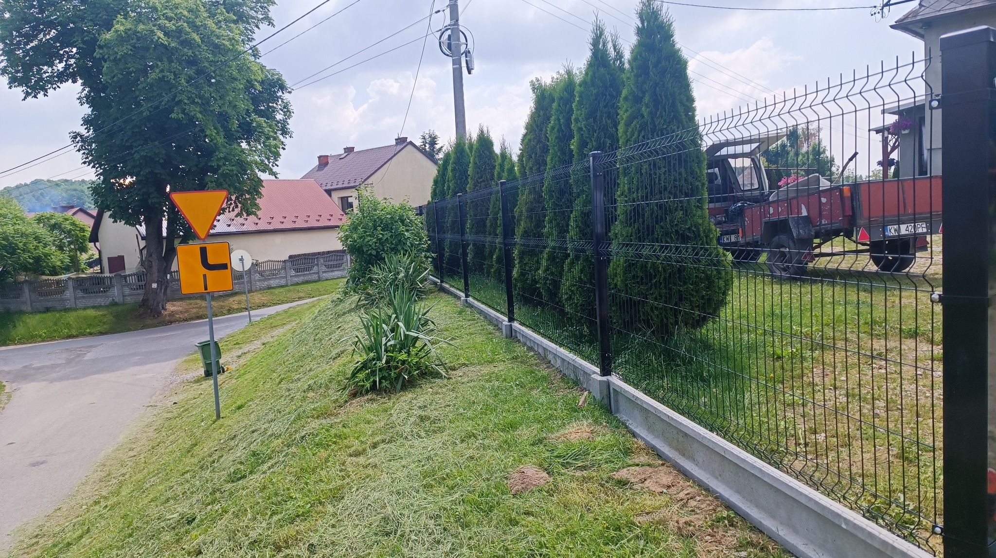 Czarny ogrodzenie panelowe na betonowej podmurówce, widok z boku, za ogrodzeniem rząd tuj i czerwony traktor z przyczepą, w tle domy.