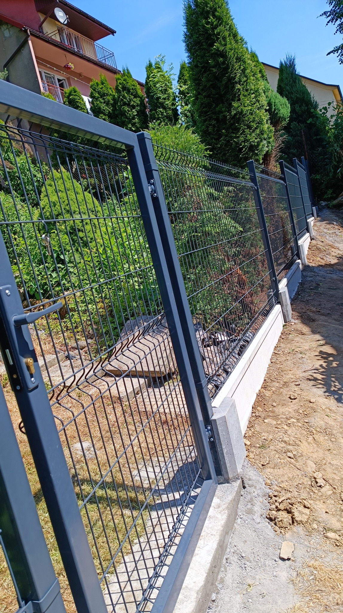 Nowoczesne ogrodzenie panelowe z metalową bramką, osadzone na betonowej podmurówce, widoczne z perspektywy bliskiej ziemi, z zielenią w tle.