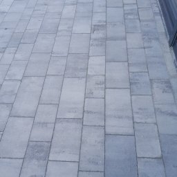 ROM-BRUK Usługi Brukarskie i Budowlane Gwóźdź Roman - Świeżo ułożona, szara kostka brukowa prostokątna tworząca chodnik przed budynkiem z widocznymi przeszkleniami.