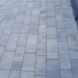 Świeżo ułożona, szara kostka brukowa prostokątna tworząca chodnik przed budynkiem z widocznymi przeszkleniami.