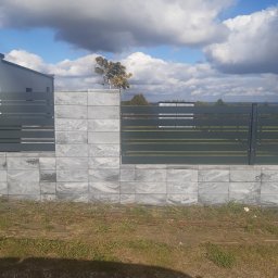 ROM-BRUK Usługi Brukarskie i Budowlane Gwóźdź Roman - Nowoczesne ogrodzenie panelowe w kolorze grafitowym, osadzone na fundamencie z szarej kostki imitującej kamień, na tle błękitnego nieba z kłębiastymi chmurami.