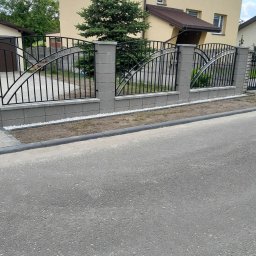 ROM-BRUK Usługi Brukarskie i Budowlane Gwóźdź Roman - Nowoczesne ogrodzenie z czarnych prętów i szarych betonowych słupków z ozdobnymi łukami, białe kamyczki dekoracyjne u podstawy, żółty dom w tle.