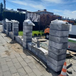 ROM-BRUK Usługi Brukarskie i Budowlane Gwóźdź Roman - Wznoszenie ogrodzenia z szarych, betonowych bloczków; widoczne słupki i fragment muru w trakcie budowy, zabezpieczone folią, pomarańczowy pachołek ostrzegawczy, paleta z materiałem budowlanym,...