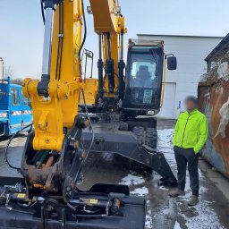 ROM-BRUK Usługi Brukarskie i Budowlane Gwóźdź Roman - Żółto-czarna koparko-ładowarka JCB na placu budowy, obok stoi mężczyzna w neonowej kurtce, w tle widoczny niebieski samochód dostawczy i blaszana ściana.
