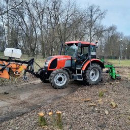 ROM-BRUK Usługi Brukarskie i Budowlane Gwóźdź Roman - Czerwono-szary traktor Zetor z zamontowanymi narzędziami do pielęgnacji terenu, w tym szczotką i frezem do pni, stoi na ubitej ziemi z widocznymi ściętymi pniami drzew, na tle parku z drzewami bez...