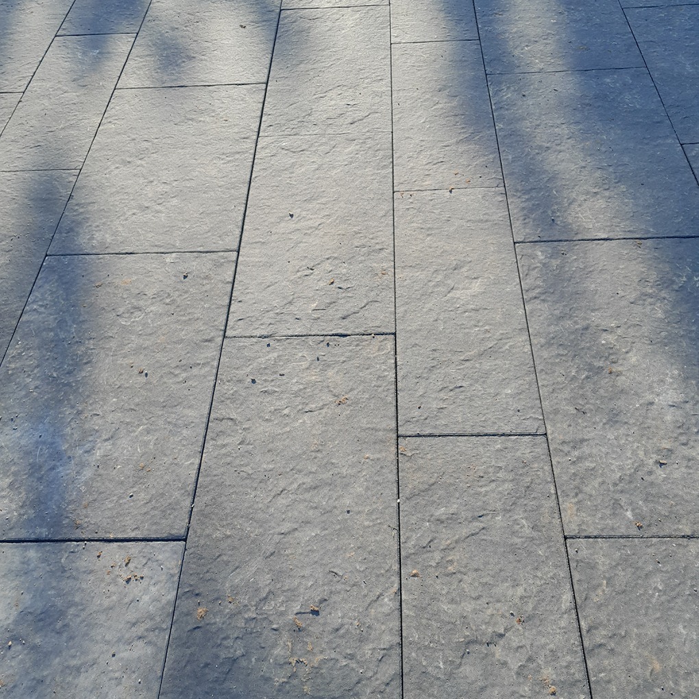 Ułożona nawierzchnia z szarych, prostokątnych płyt kamiennych z widocznymi cieniami drzew i drobnymi zabrudzeniami.