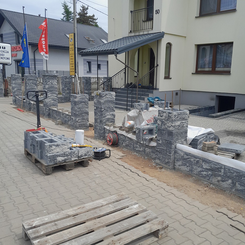 Budowa ogrodzenia z szarej, łupanej kostki betonowej. Widoczne słupki w trakcie wznoszenia, paleta z pustakami, narzędzia i budynek mieszkalny w tle.