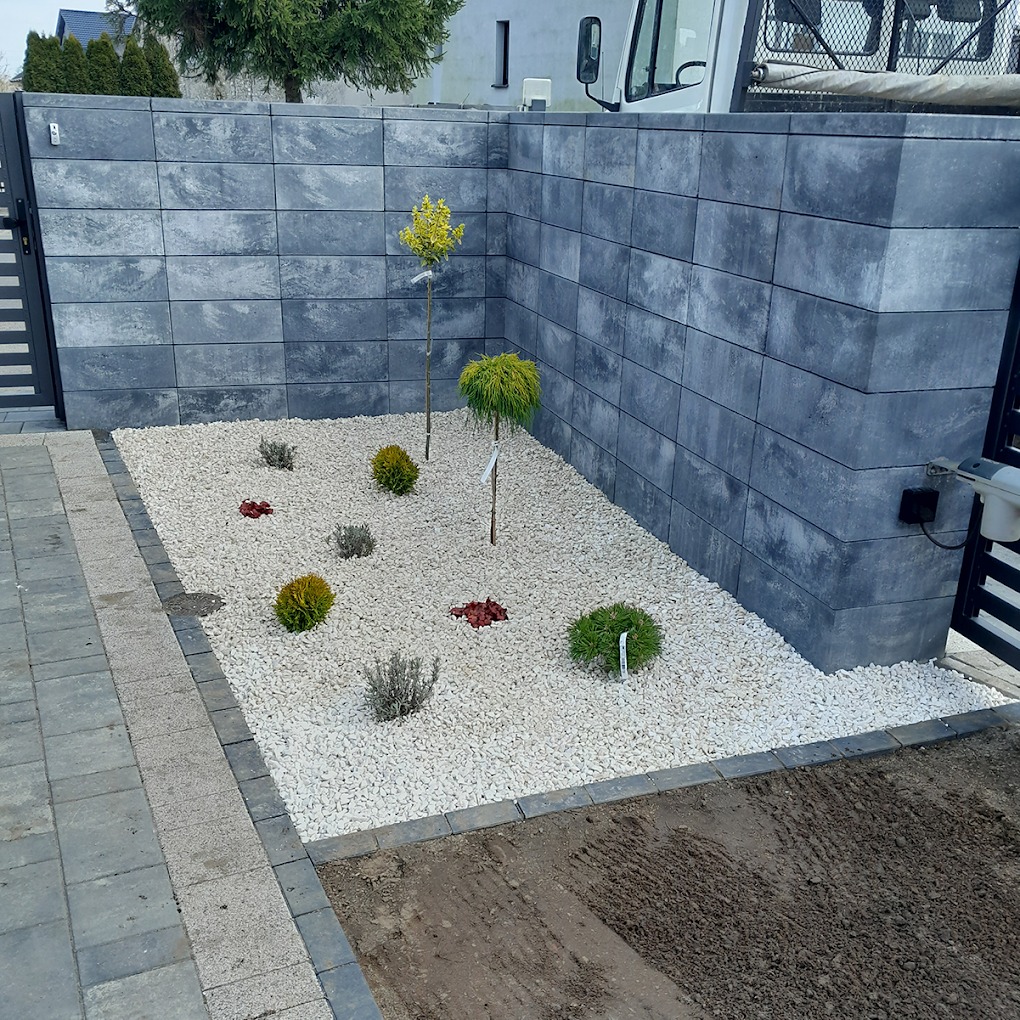 Nowoczesny ogród z szarego kamienia, niskimi krzewami i młodymi drzewkami, otoczony murem z betonowych bloków i fragmentem ogrodzenia z automatem.