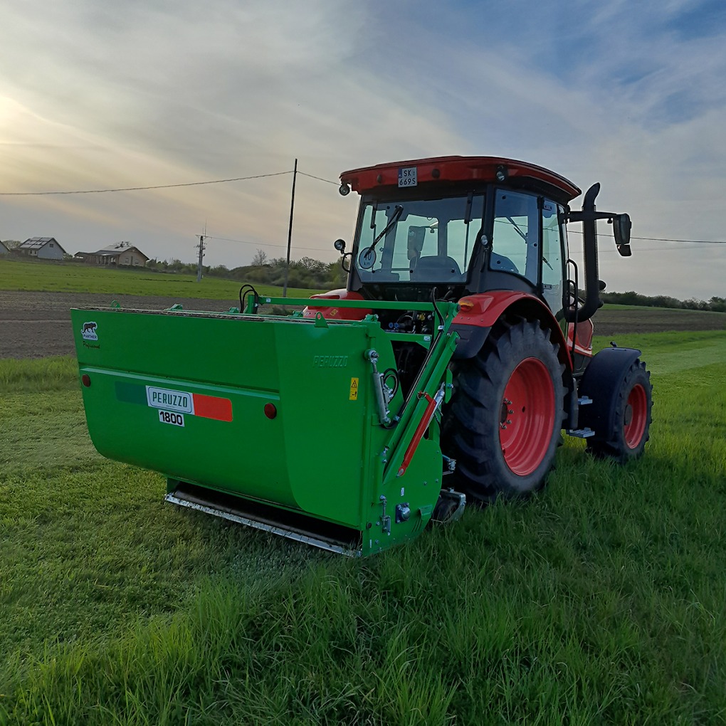 Czerwony traktor z zielonym kosiarką Peruzzo 1800 na trawiastym polu, w tle zabudowania gospodarcze i zachodzące słońce.