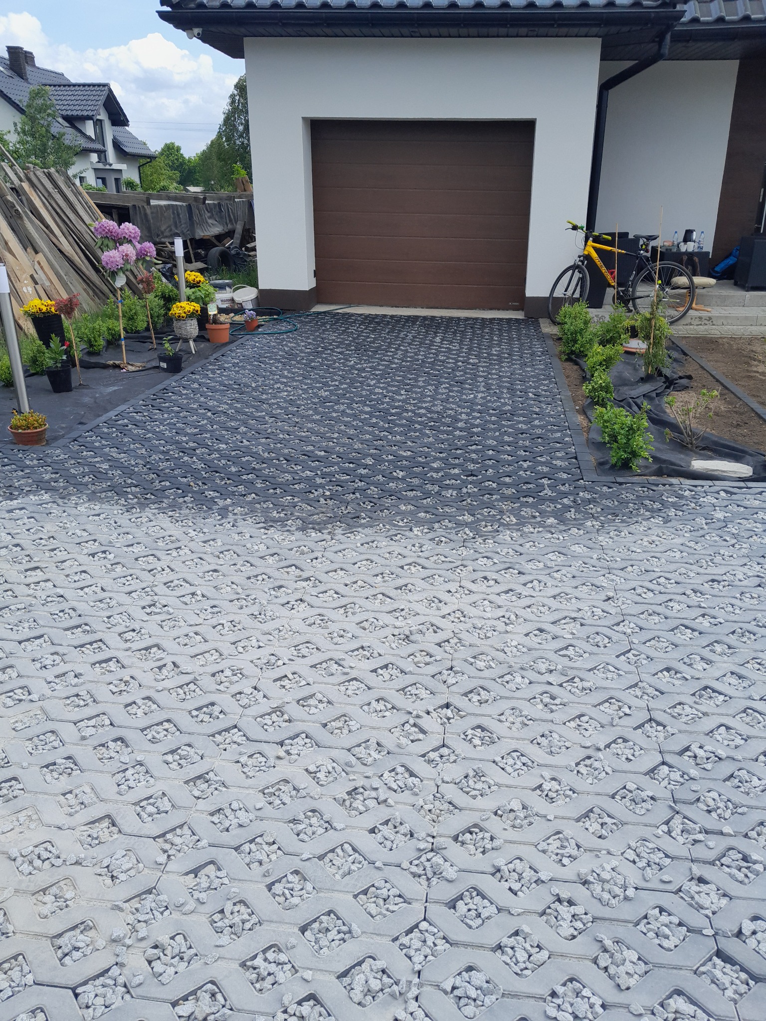 Częściowo ułożony podjazd z ażurowych płyt betonowych wypełnionych drobnym grysem, widok z góry, na dalszym planie garaż, rower i przygotowane sadzonki.