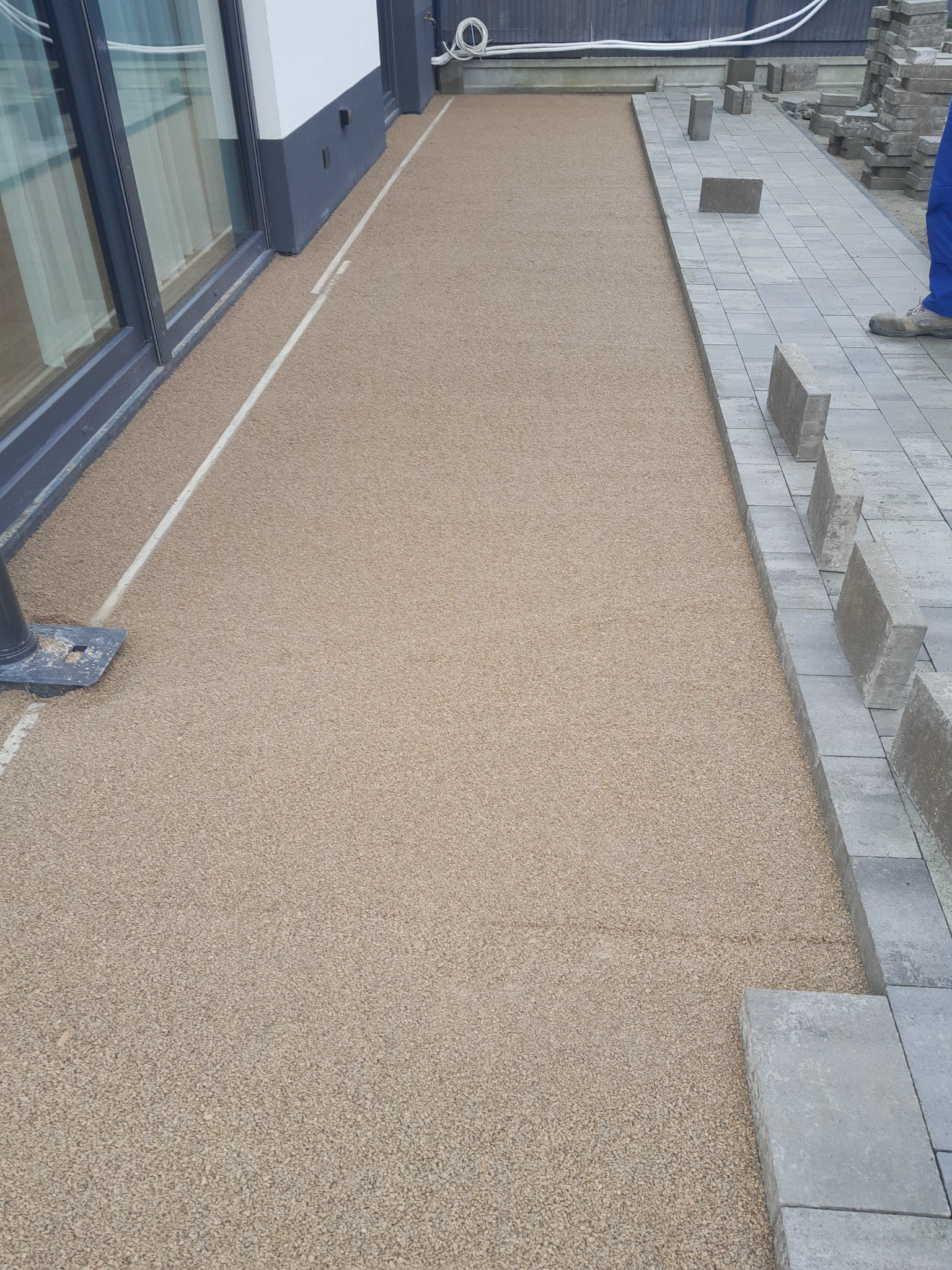 Przygotowane podłoże z piasku kwarcowego pod układanie szarej kostki brukowej przed budynkiem z dużymi oknami.