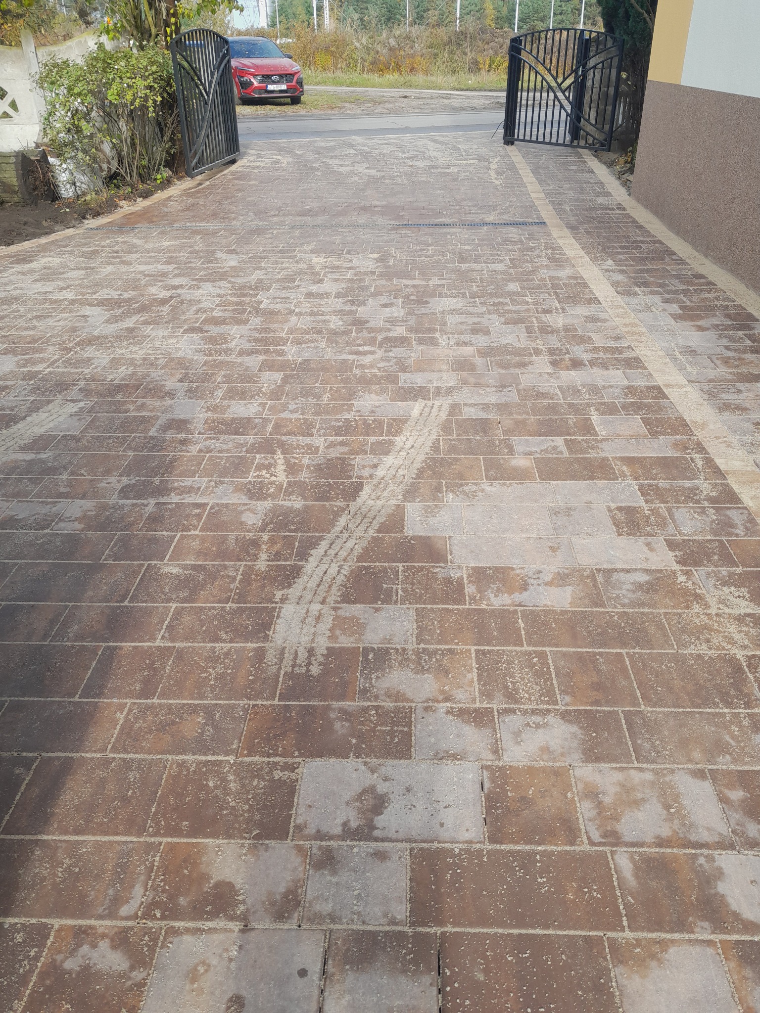 Nowa, brązowa nawierzchnia z kostki brukowej ułożona na podjeździe, z widocznymi śladami opon, prowadząca do metalowej bramy wjazdowej i czerwonego samochodu w tle.