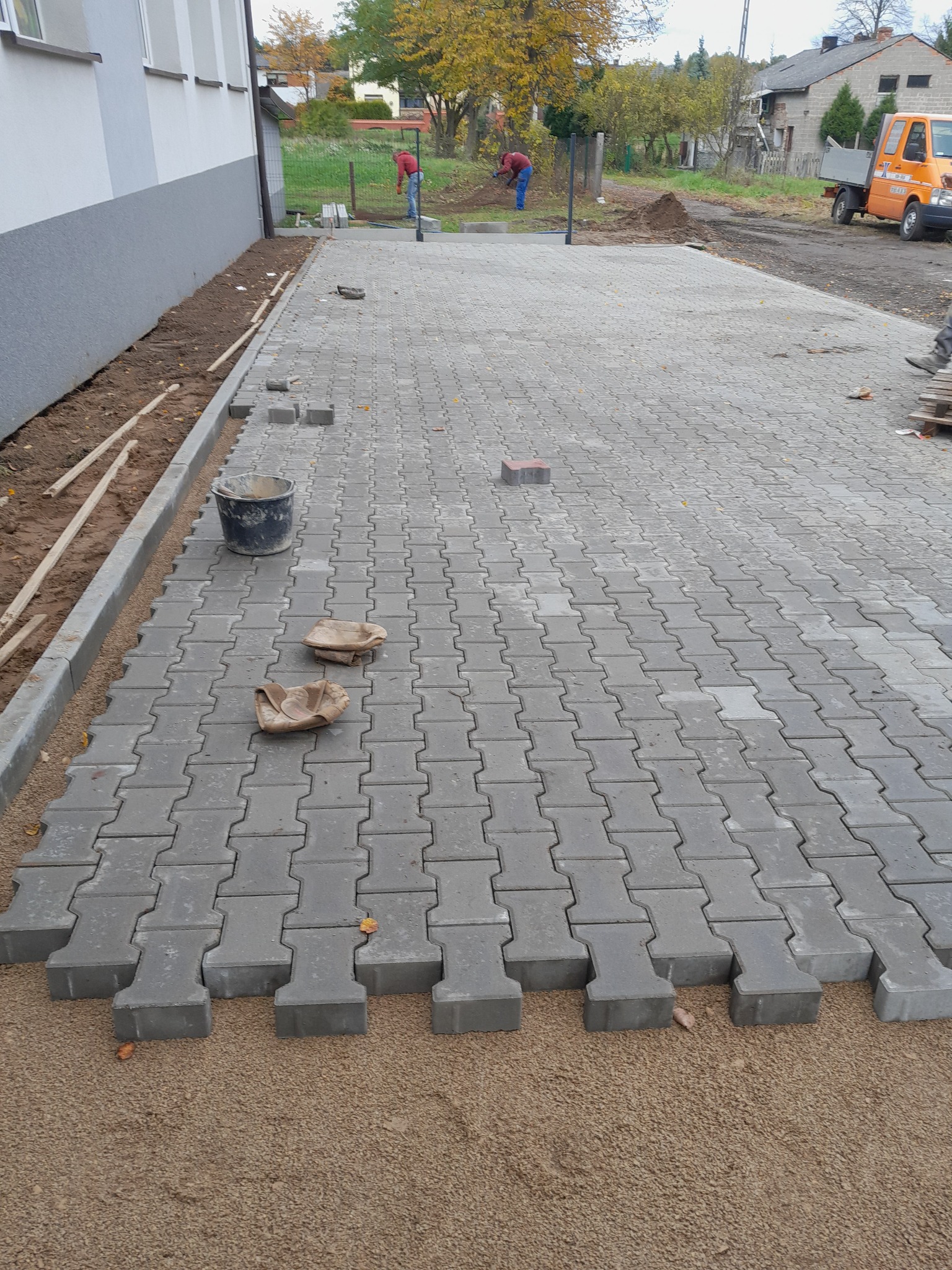 Układanie szarej kostki brukowej typu I na podsypce piaskowej, widoczne rękawice robocze, wiadro i fragmenty niezabudowane, w tle pracownicy i samochód dostawczy.