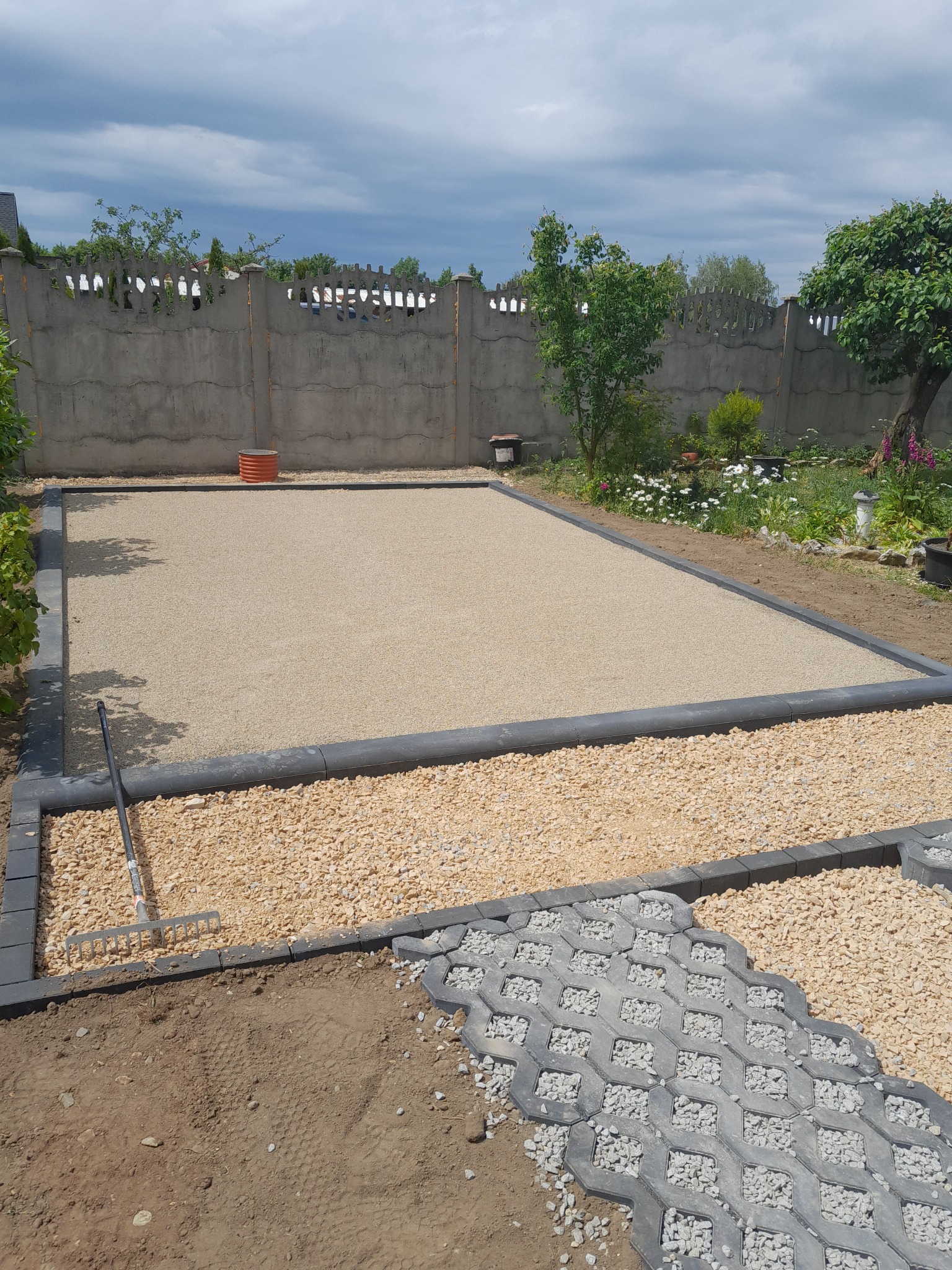 Przygotowany teren pod budowę boiska do gry w bule z obramowaniem z betonowych obrzeży i częściowo ułożoną ażurową nawierzchnią wypełnioną drobnym kruszywem, w tle betonowy płot i zieleń.