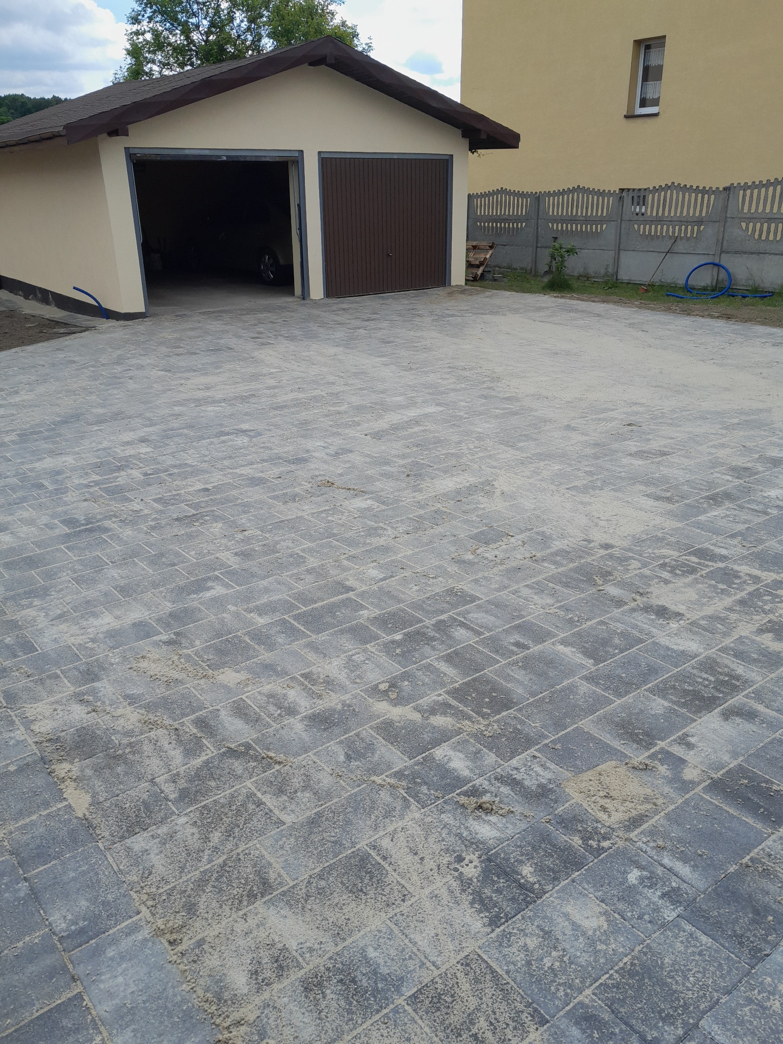 Nowo ułożona, szara kostka brukowa przed garażem z częściowo otwartymi, brązowymi drzwiami, z widocznymi śladami piasku na powierzchni i betonowym ogrodzeniem w tle.