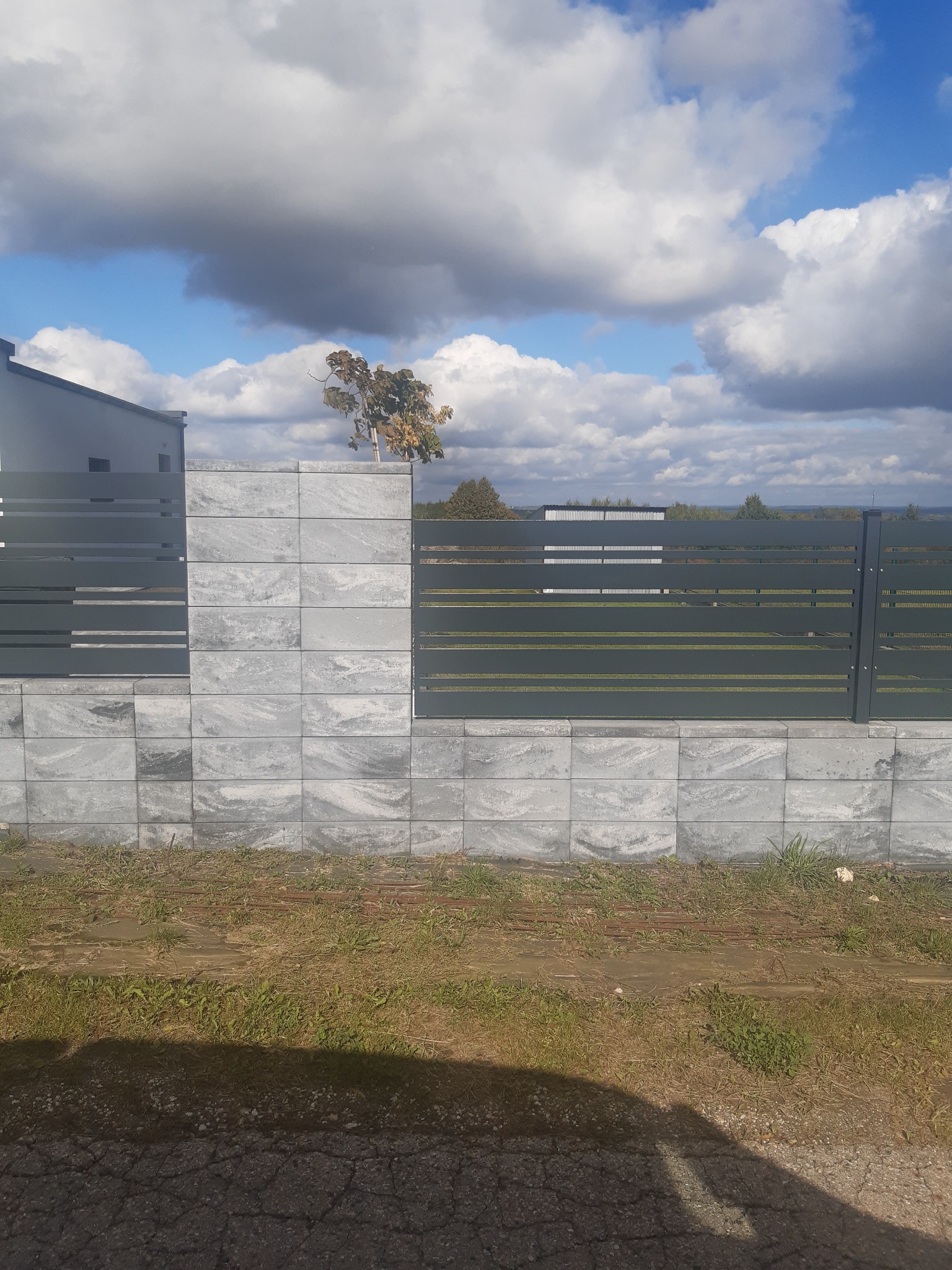 Nowoczesne ogrodzenie panelowe w kolorze grafitowym, osadzone na fundamencie z szarej kostki imitującej kamień, na tle błękitnego nieba z kłębiastymi chmurami.