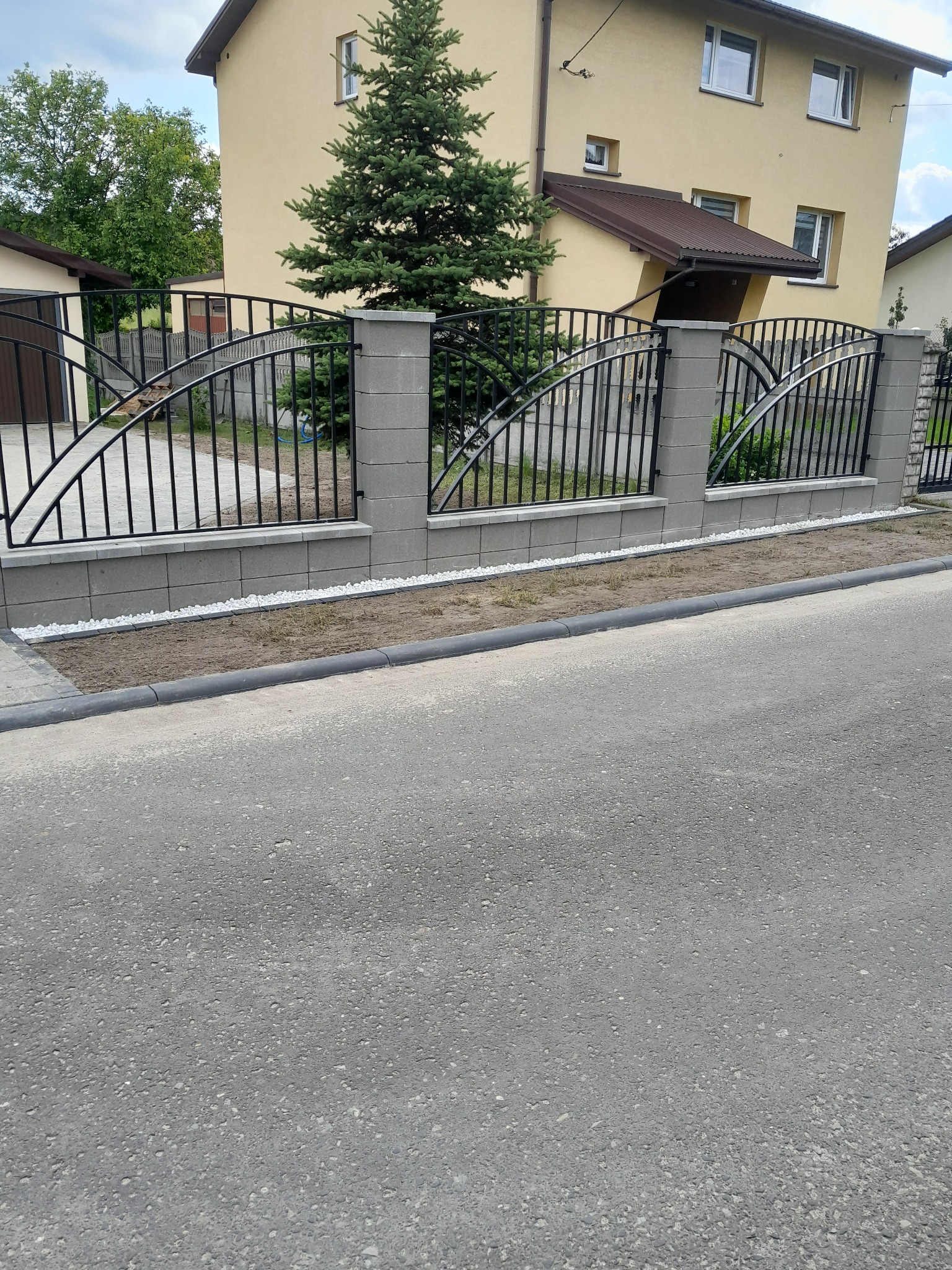 Nowoczesne ogrodzenie z czarnych prętów i szarych betonowych słupków z ozdobnymi łukami, białe kamyczki dekoracyjne u podstawy, żółty dom w tle.