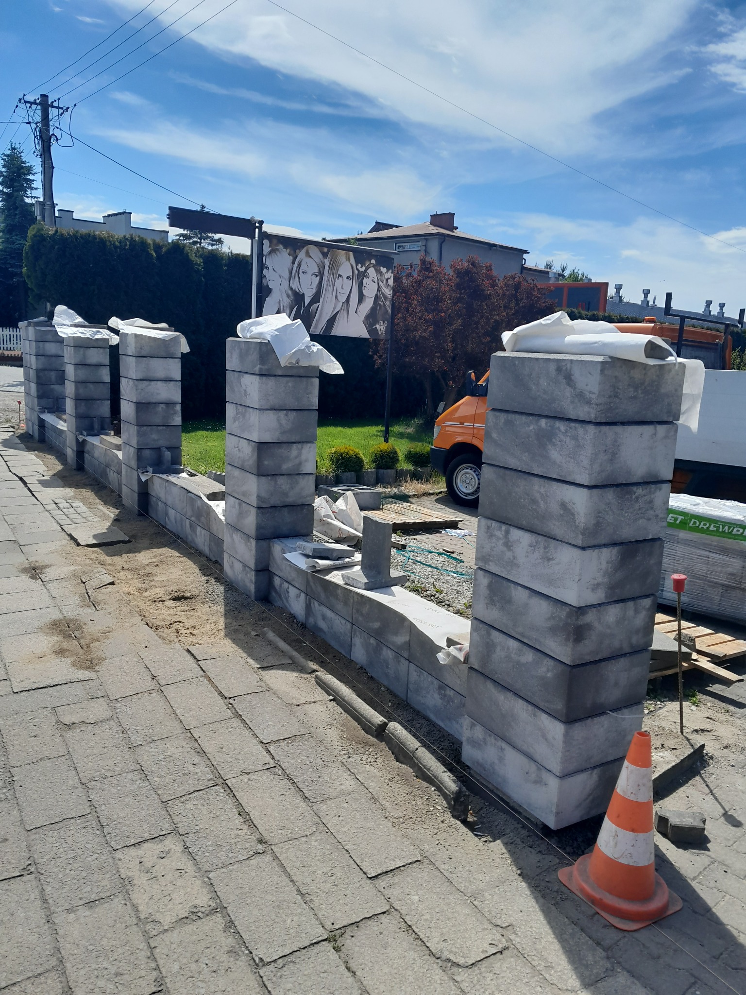 Wznoszenie ogrodzenia z szarych, betonowych bloczków; widoczne słupki i fragment muru w trakcie budowy, zabezpieczone folią, pomarańczowy pachołek ostrzegawczy, paleta z materiałem budowlanym,...