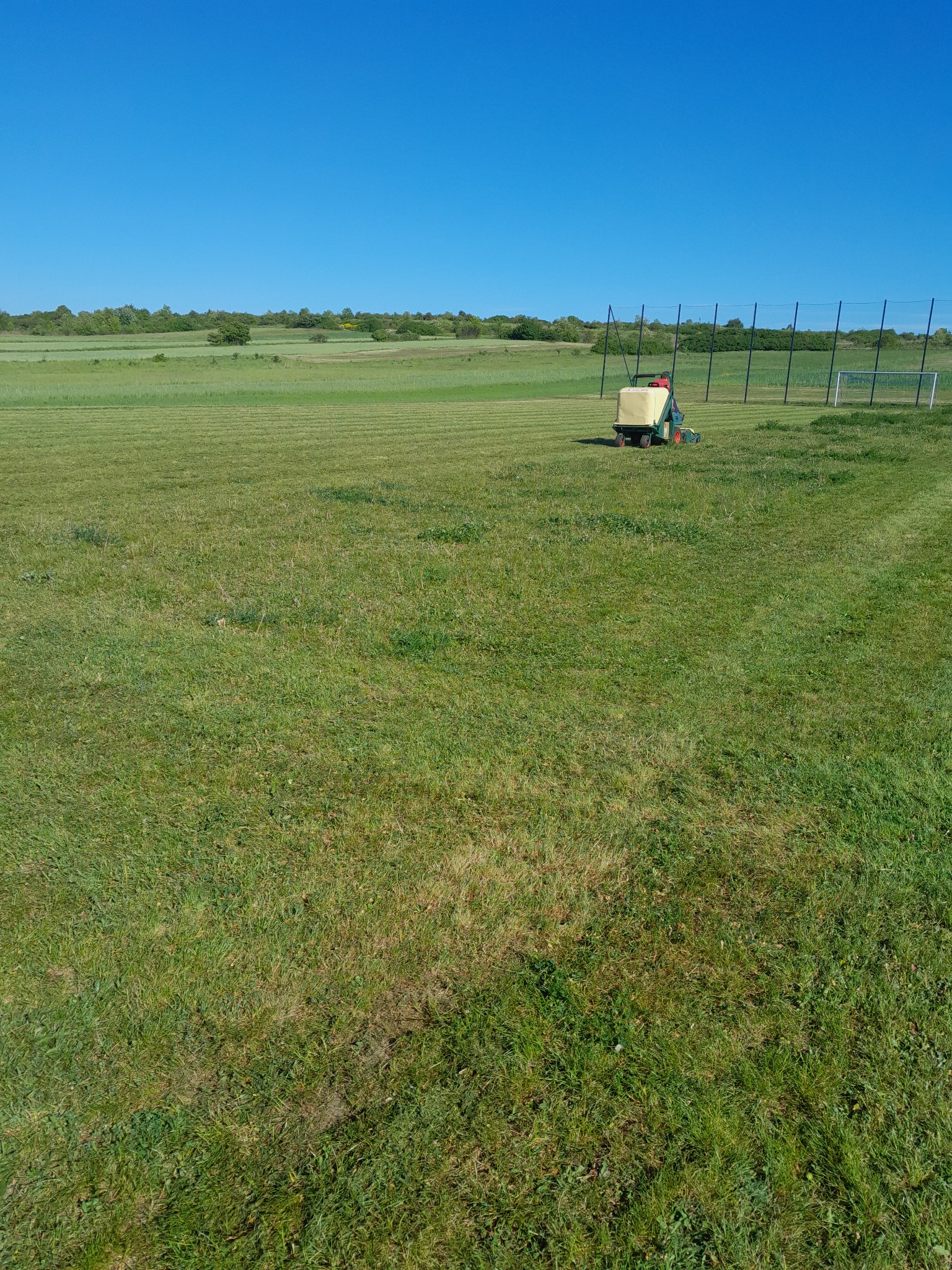 Traktor z kosiarką w trakcie pracy na rozległym, zielonym boisku sportowym z bramkami w tle, widok w słoneczny dzień.
