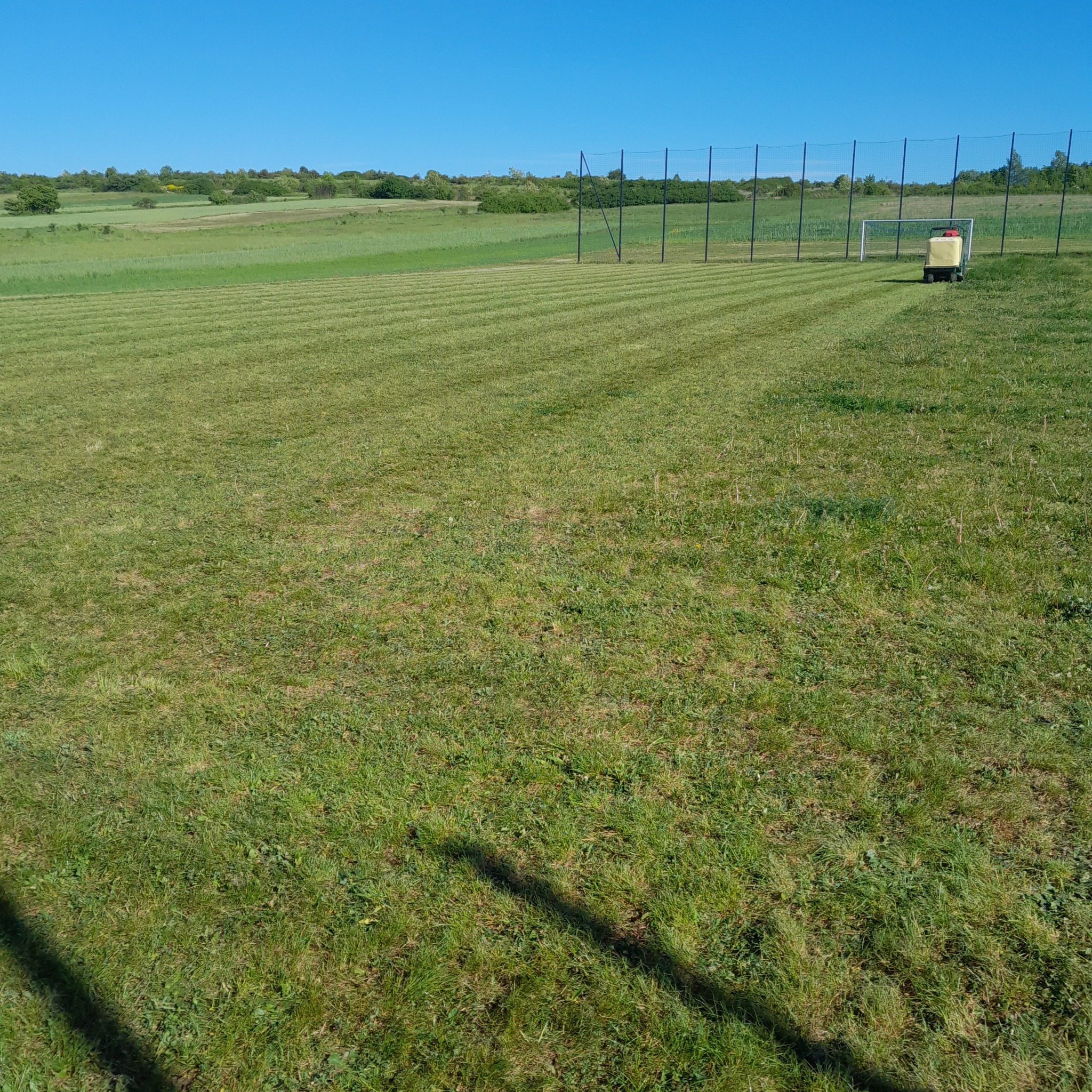 Szeroki kadr na równo przystrzyżoną trawę na boisku sportowym z widocznymi pasami po kosiarce, w tle siatka i bramka oraz maszyna do malowania linii, błękitne niebo