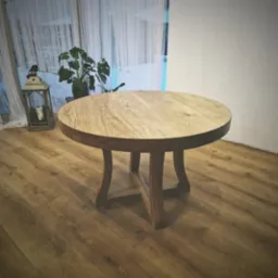 Okrągły stół z jasnego drewna z ciemniejszym obrzeżem, stojący na drewnianej podłodze, z dekoracyjną latarnią i rośliną w tle przy oknie z białymi firankami.