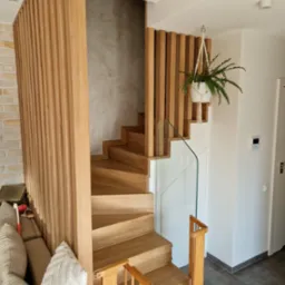 Nowoczesne schody z drewna z ażurową balustradą z pionowych listew i szklanym panelem, uzupełnione wiszącą rośliną w białej doniczce.