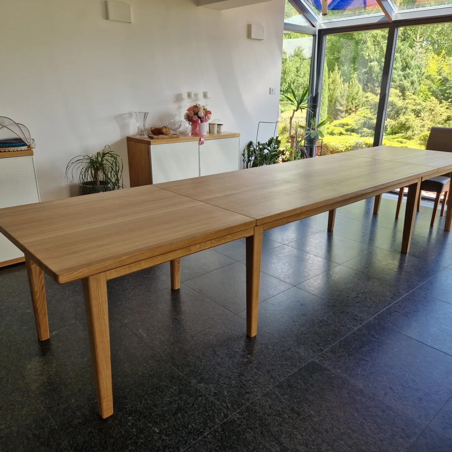 Długi, rozkładany stół z jasnego drewna w nowoczesnej jadalni z dużym oknem i widokiem na ogród. Stół stoi na ciemnej, błyszczącej podłodze.