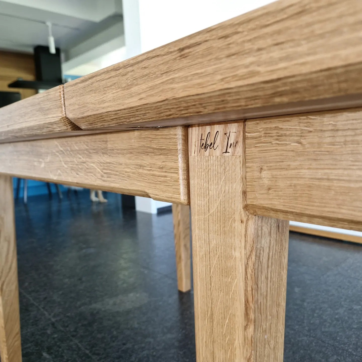 Fragment dębowego stołu z widocznym połączeniem blatu i nogi, na której wygrawerowano logo producenta.
