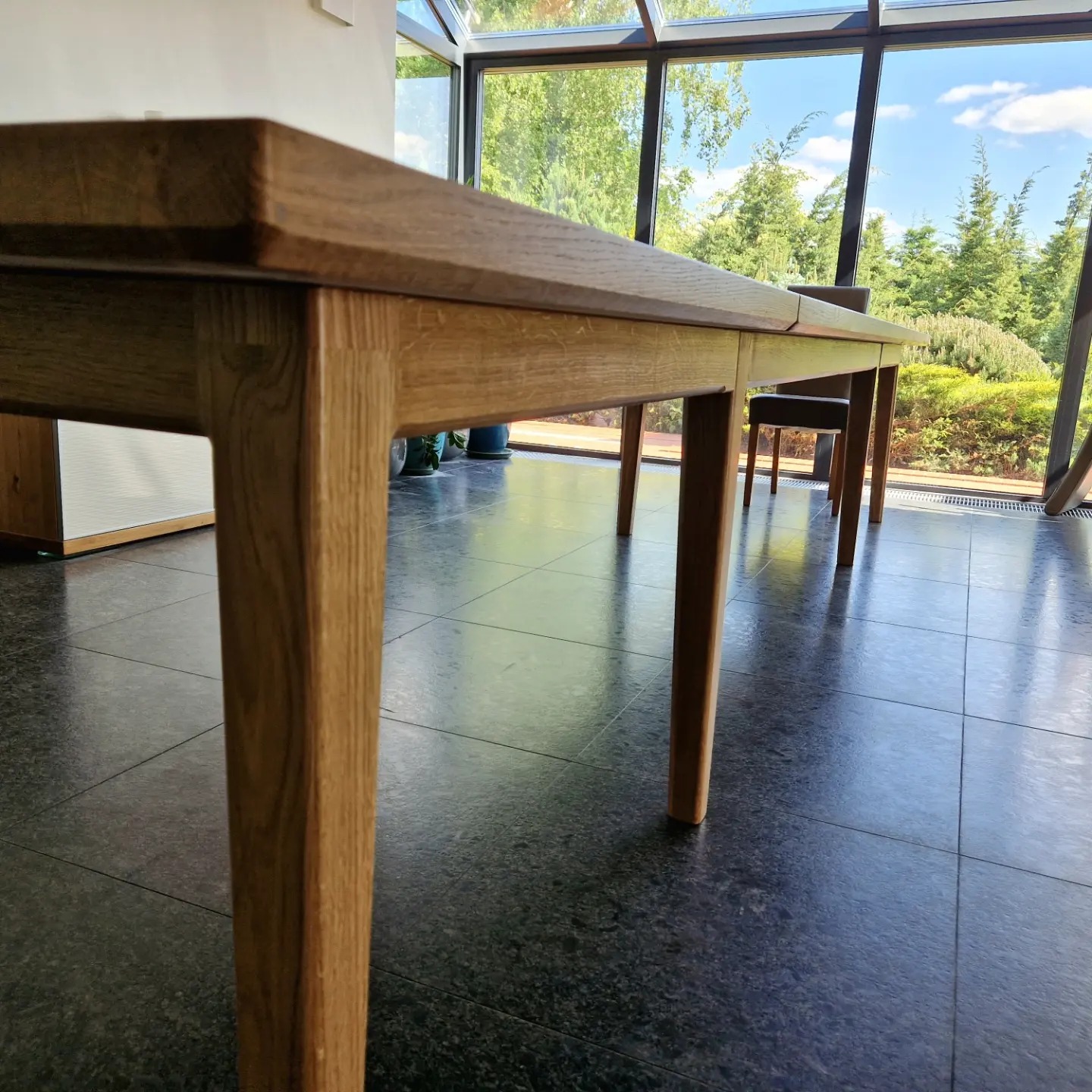 Rozłożony drewniany stół w jasnym pomieszczeniu z dużymi oknami, widok z niskiego kąta, ukazujący strukturę drewna i łączenie blatu.