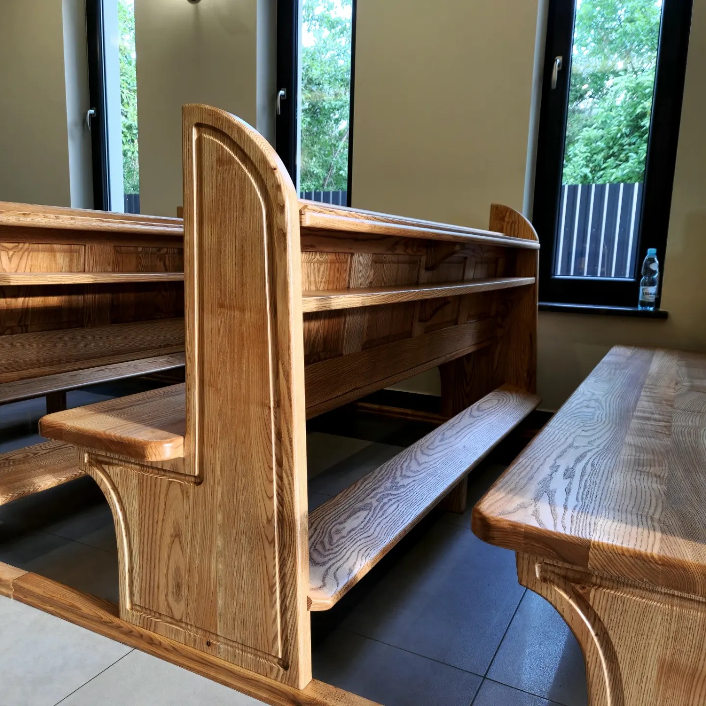 Jasne, drewniane ławki kościelne z oparciami i siedziskami o wyraźnym usłojeniu, ustawione w rzędzie przy oknach z widokiem na zieleń.