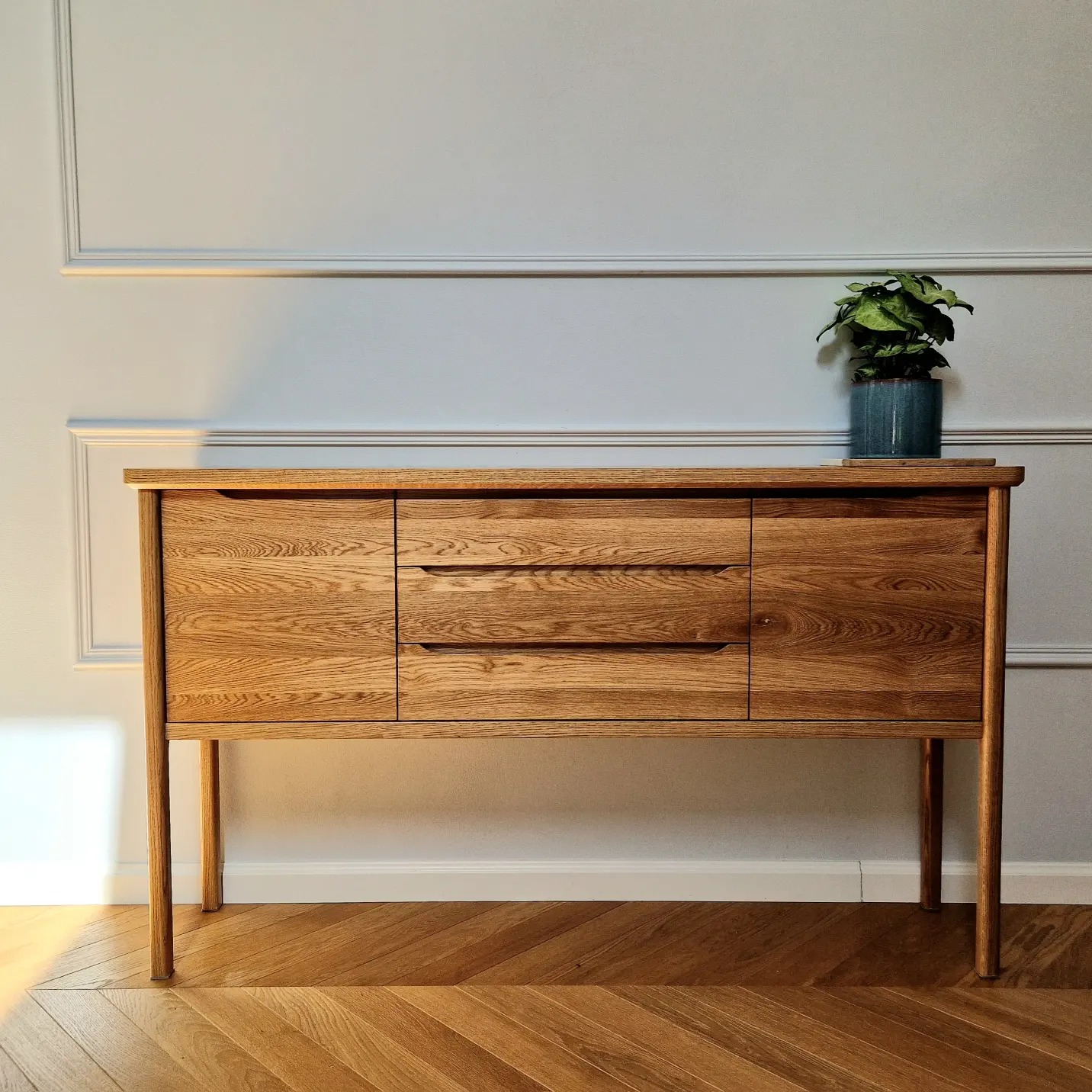 Dębowa komoda z trzema szufladami i szafkami, stojąca na drewnianym parkiecie w jodełkę, z rośliną doniczkową na blacie.
