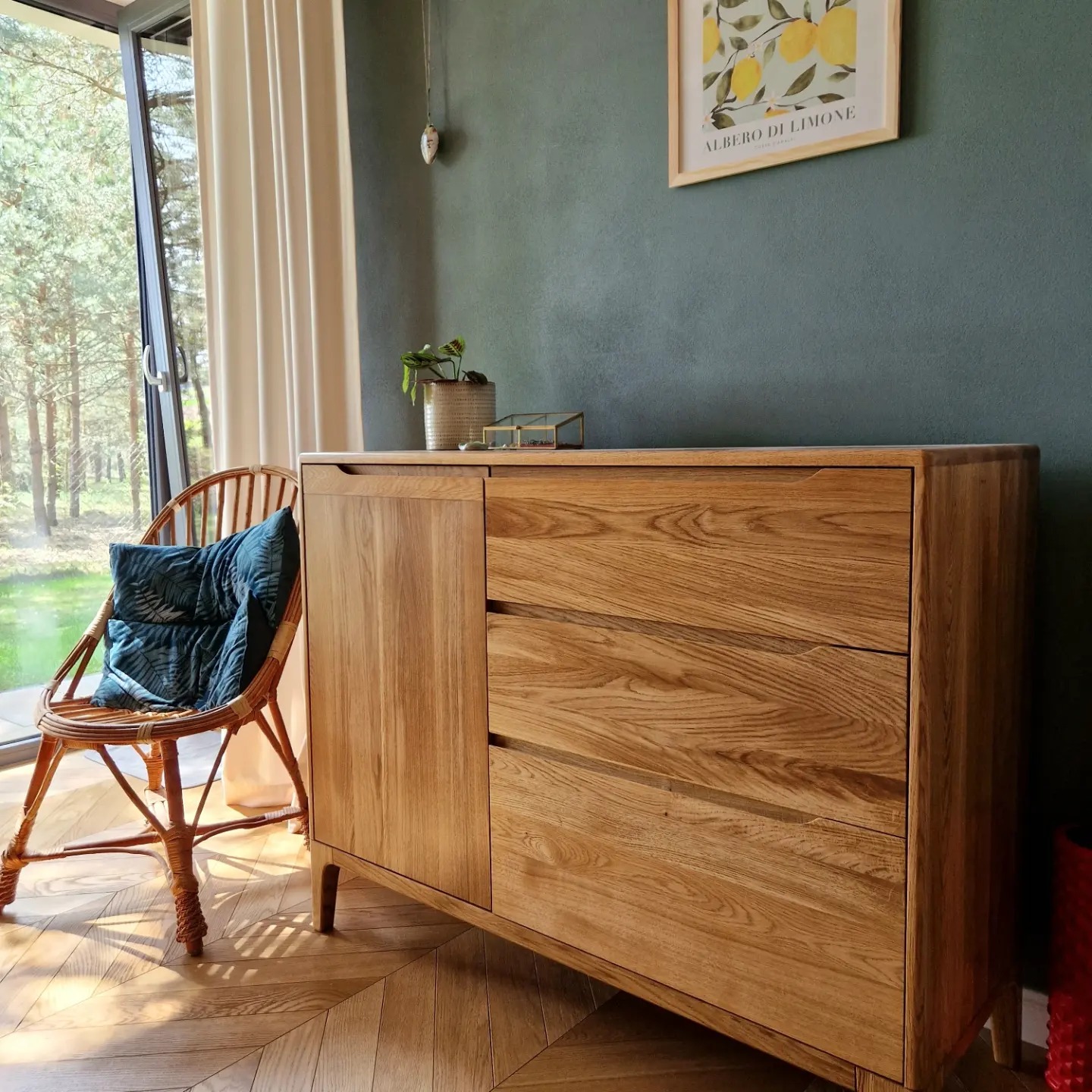 Komoda z jasnego drewna z trzema szufladami i szafką, stojąca na parkiecie w jodełkę, obok wiklinowego fotela z poduszką, na tle ciemnozielonej ściany z obrazkiem.