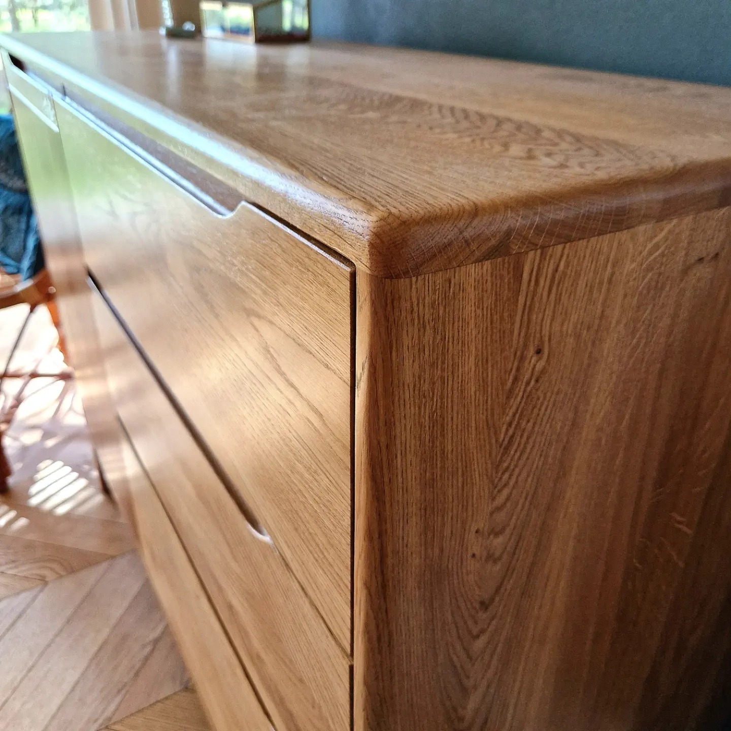 Detal prezentujący wykończenie komody z jasnego drewna dębowego, z frezowanymi uchwytami szuflad i zaokrąglonymi krawędziami blatu, na tle parkietu w jodełkę.