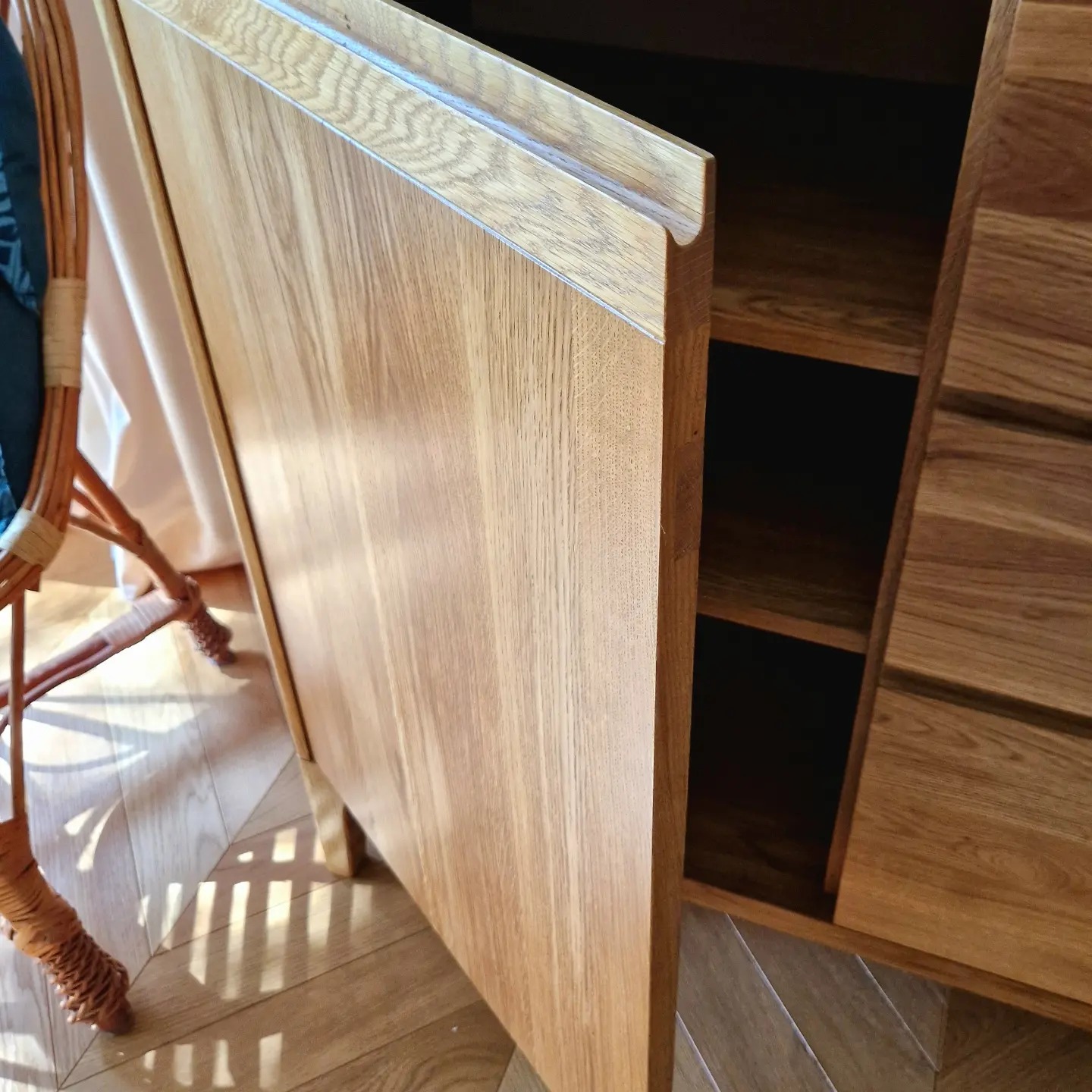 Otwarta szafka z jasnego drewna dębowego z półkami, widoczna na tle parkietu ułożonego w jodełkę, obok krzesło z wikliny.