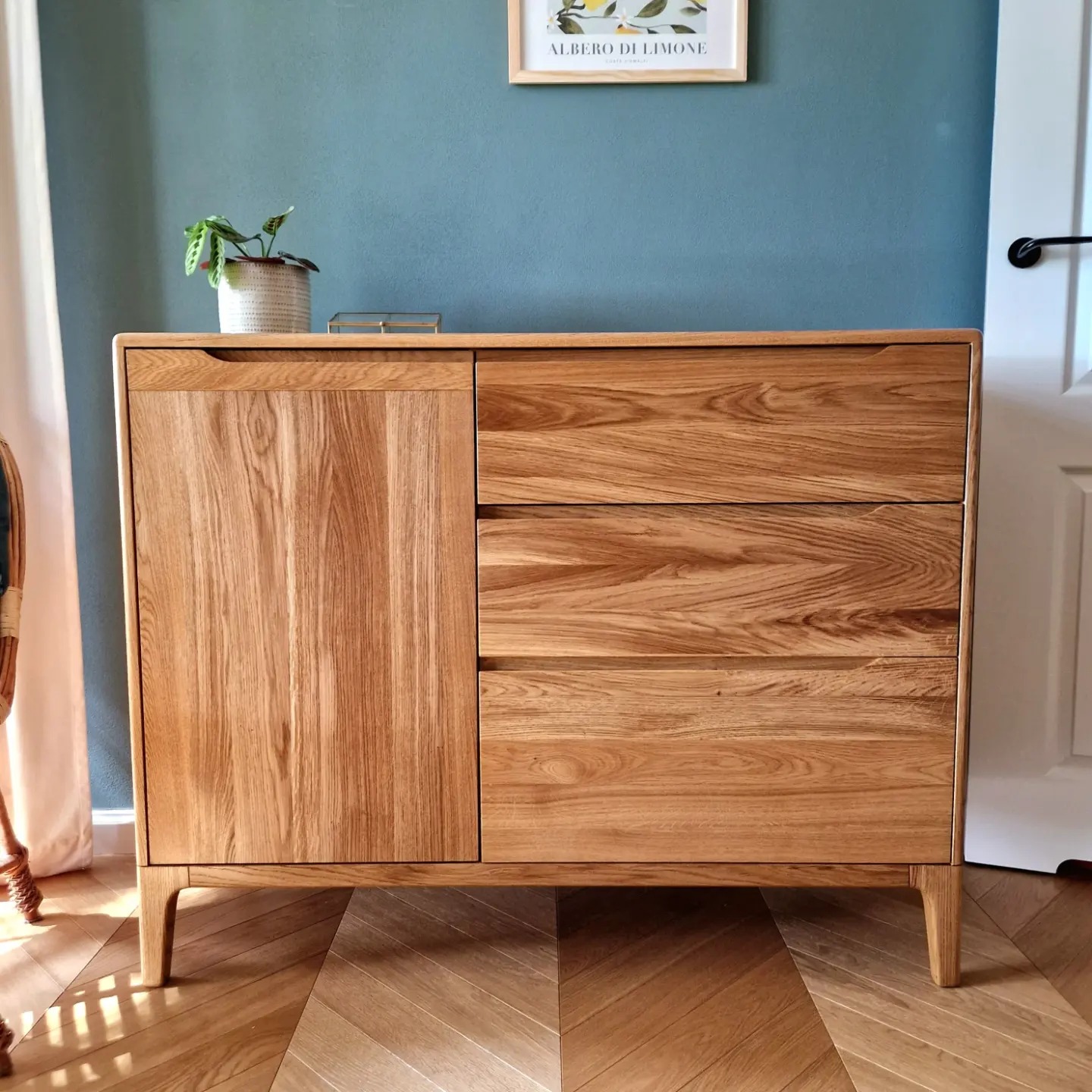 Komoda z jasnego drewna dębowego z trzema szufladami i drzwiczkami, na tle turkusowej ściany z plakatem i białych drzwi.