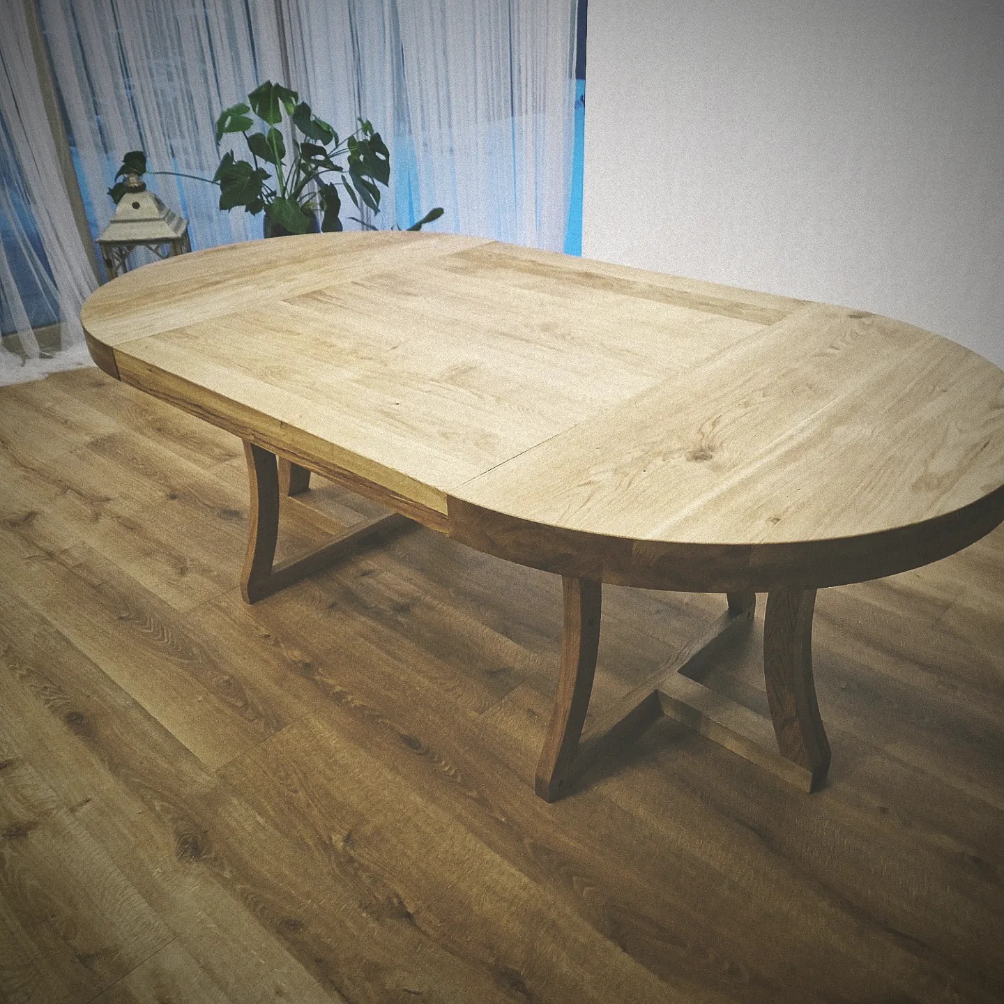 Rozkładany, owalny stół drewniany z jasnym blatem i ciemniejszymi, giętymi nogami na tle drewnianej podłogi i zasłon