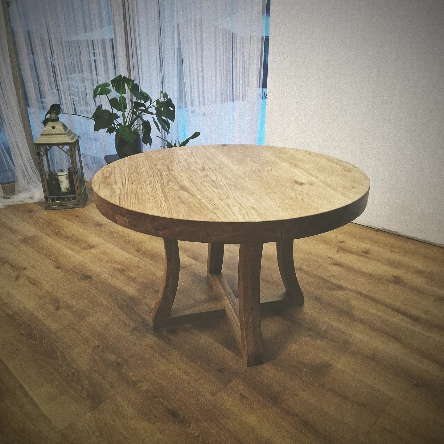 Okrągły stół z jasnego drewna z ciemniejszym obrzeżem, stojący na drewnianej podłodze, z dekoracyjną latarnią i rośliną w tle przy oknie z białymi firankami.