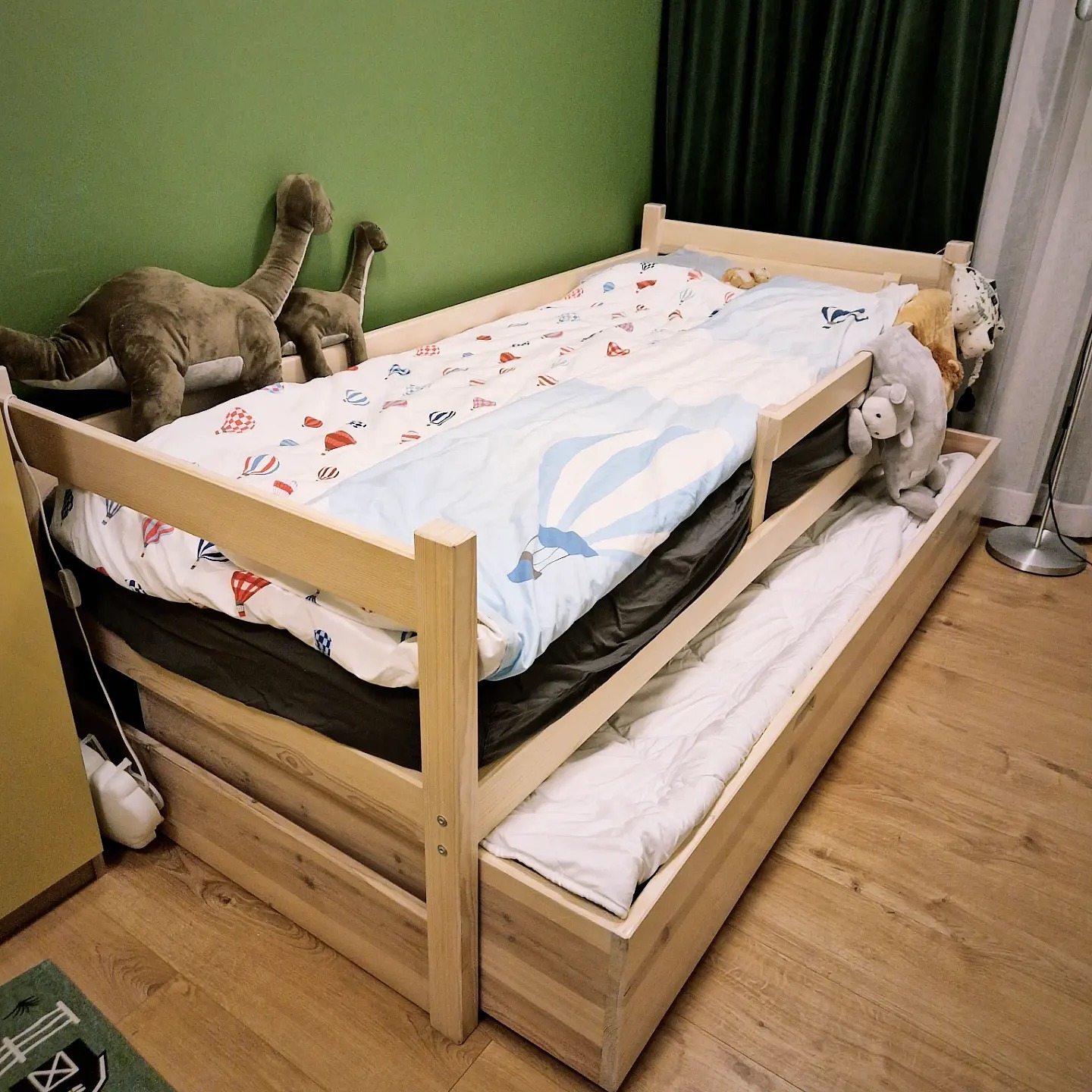 Łóżko dziecięce z drewna sosnowego z wysuwanym dodatkowym spaniem, pościel w balony i dinozaury pluszowe na zielonej ścianie.