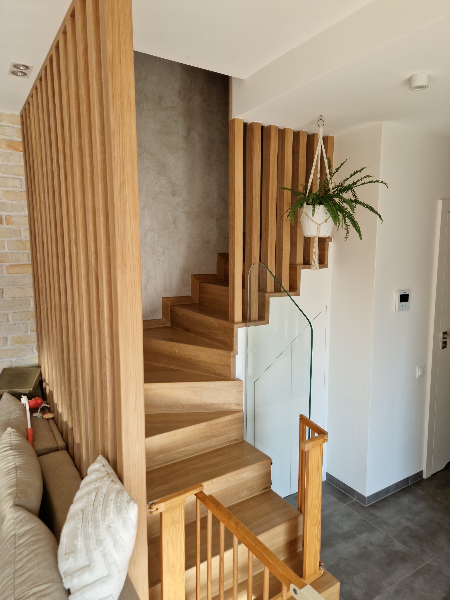 Nowoczesne schody z drewna z ażurową balustradą z pionowych listew i szklanym panelem, uzupełnione wiszącą rośliną w białej doniczce.