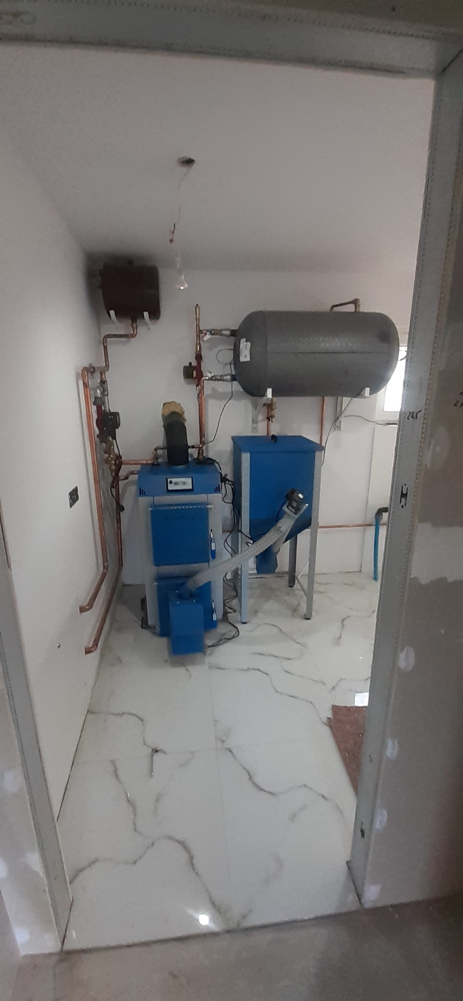 Nowoczesna kotłownia z piecem na pellet, zbiornikiem buforowym i miedzianą instalacją hydrauliczną w pomieszczeniu z białymi ścianami i marmurową podłogą, widok przez framugę drzwi.