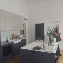 Grzegorz Pałasz - Nowoczesna kuchnia z wyspą, białe szafki górne i ciemnogranatowe dolne, biały blat, złota armatura, na wyspie owoce i dekoracje, w tle osoba rozmawiająca przez telefon.