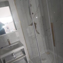 Grzegorz Pałasz - Nowoczesna łazienka z kabiną prysznicową ze złotymi elementami i umywalką nablatową, wykończona białymi płytkami imitującymi marmur, widoczne odbicie osoby w lustrze.