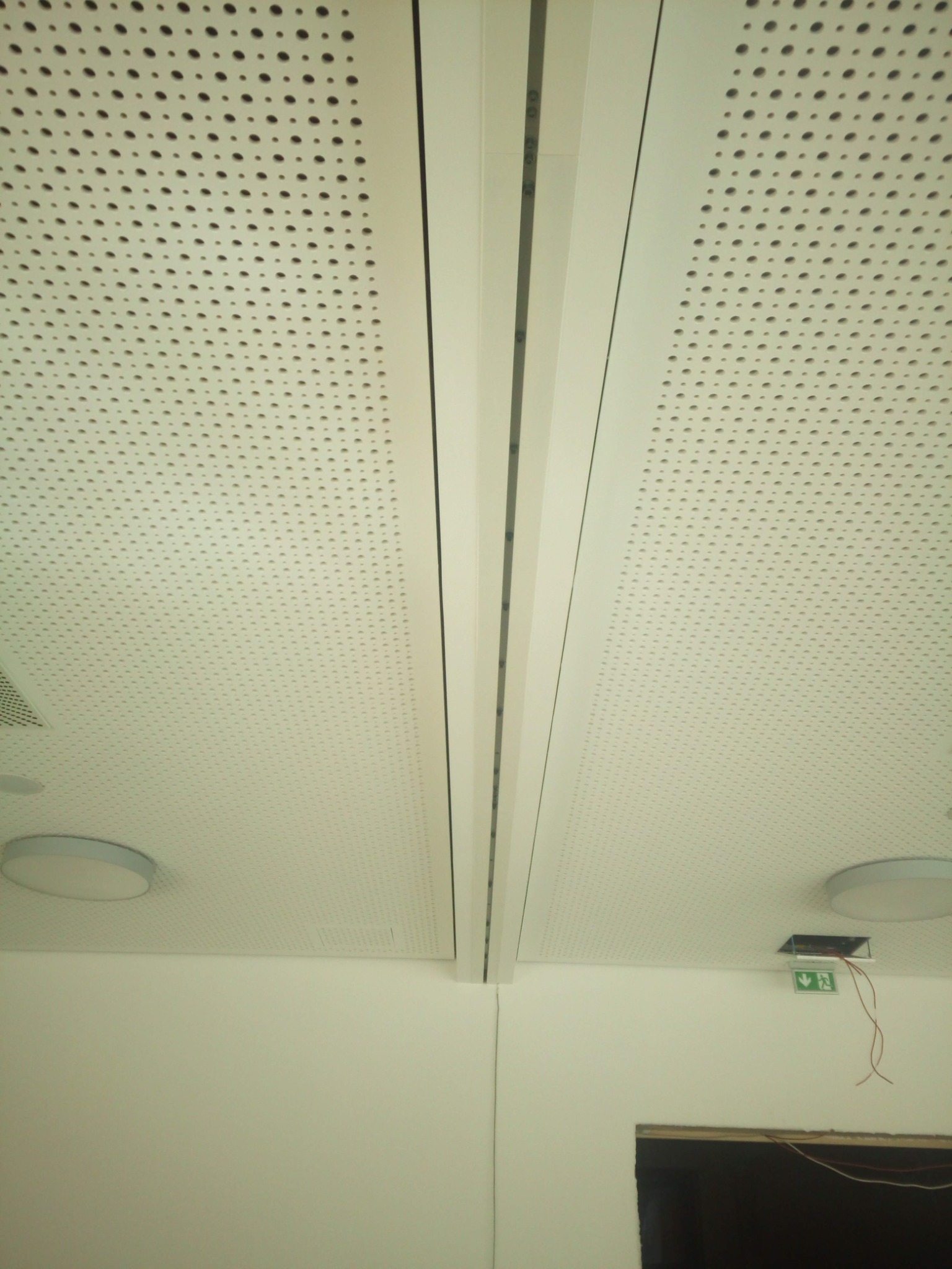 Biały sufit podwieszany z perforowanej płyty, widoczne panele i oprawy oświetleniowe, znak ewakuacyjny z wystającymi przewodami.