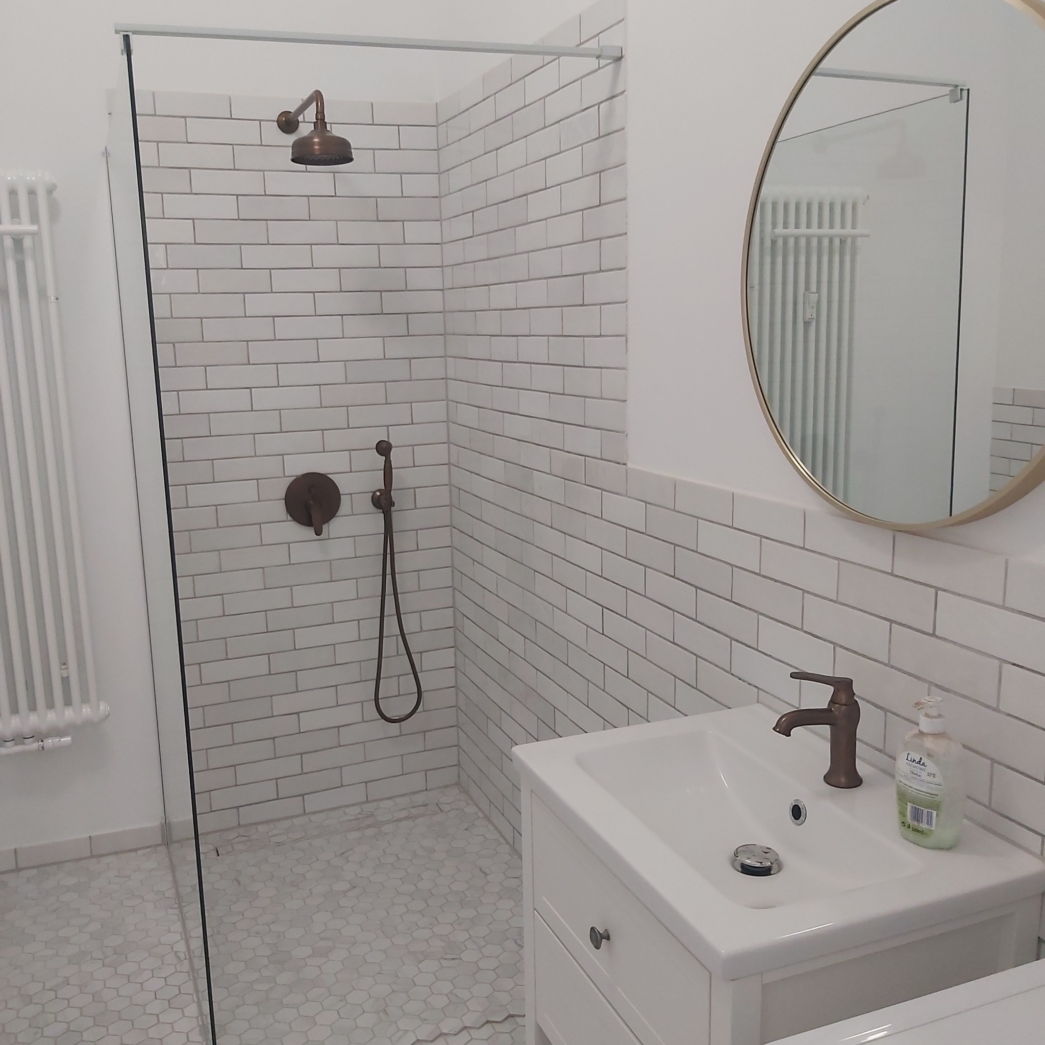 Nowoczesna łazienka z białymi, cegiełkowymi płytkami, armaturą w kolorze starego złota, kabiną prysznicową typu walk-in i okrągłym lustrem.