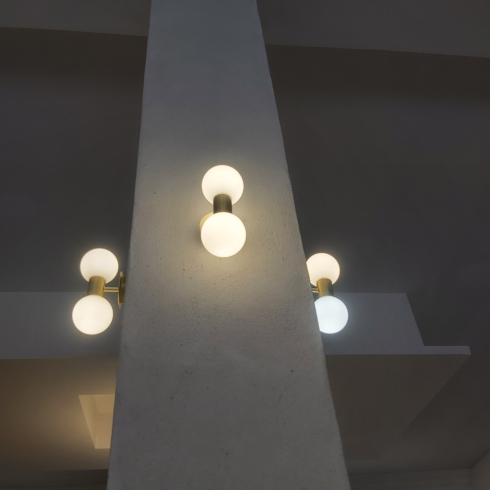 Dwie podwójne lampy kuliste z mosiężnymi uchwytami zamontowane na szarej, betonowej kolumnie w surowym wnętrzu.